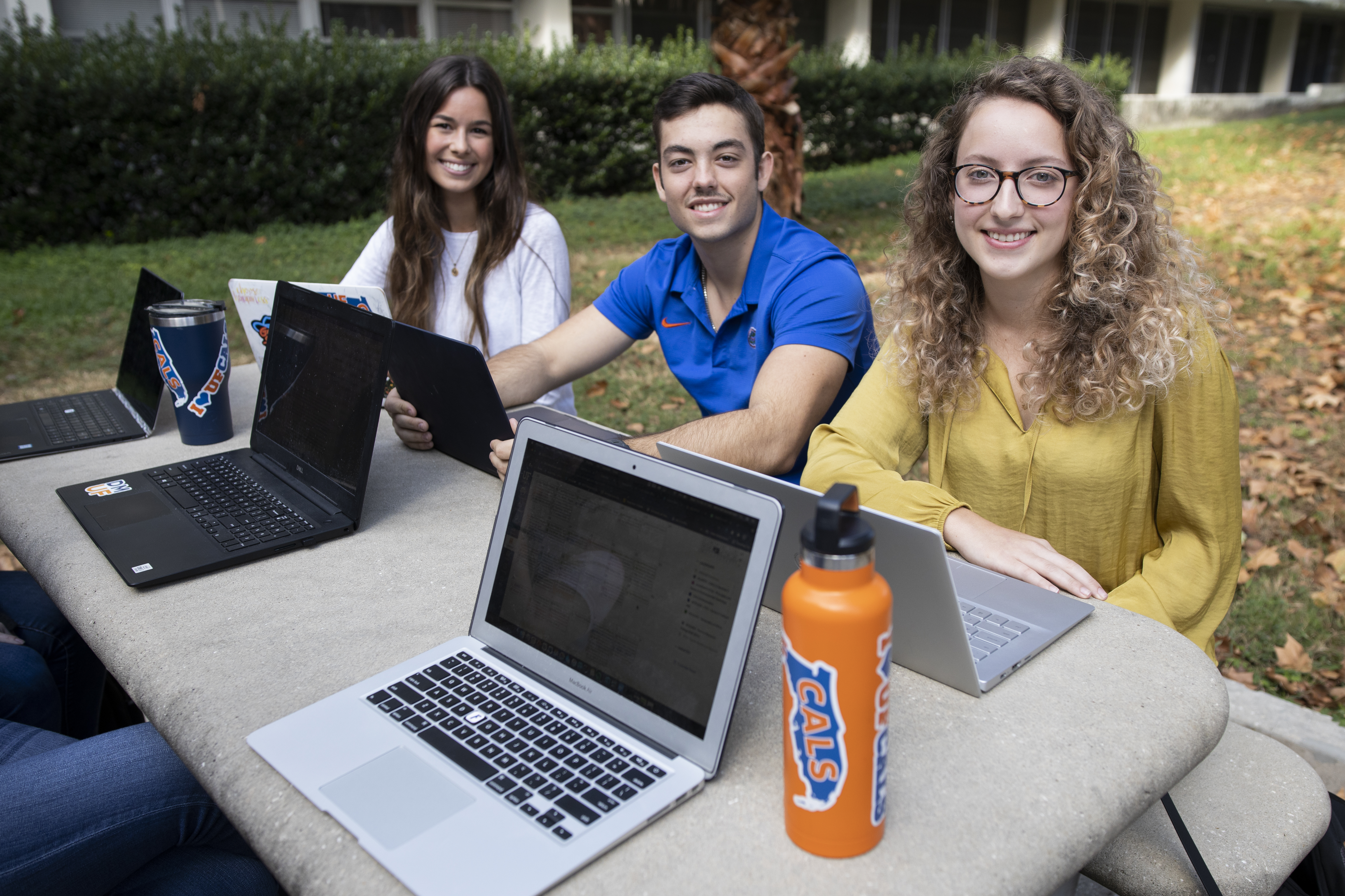 Three students in focus sitting at an outdoor concrete picnic table working on their laptops.